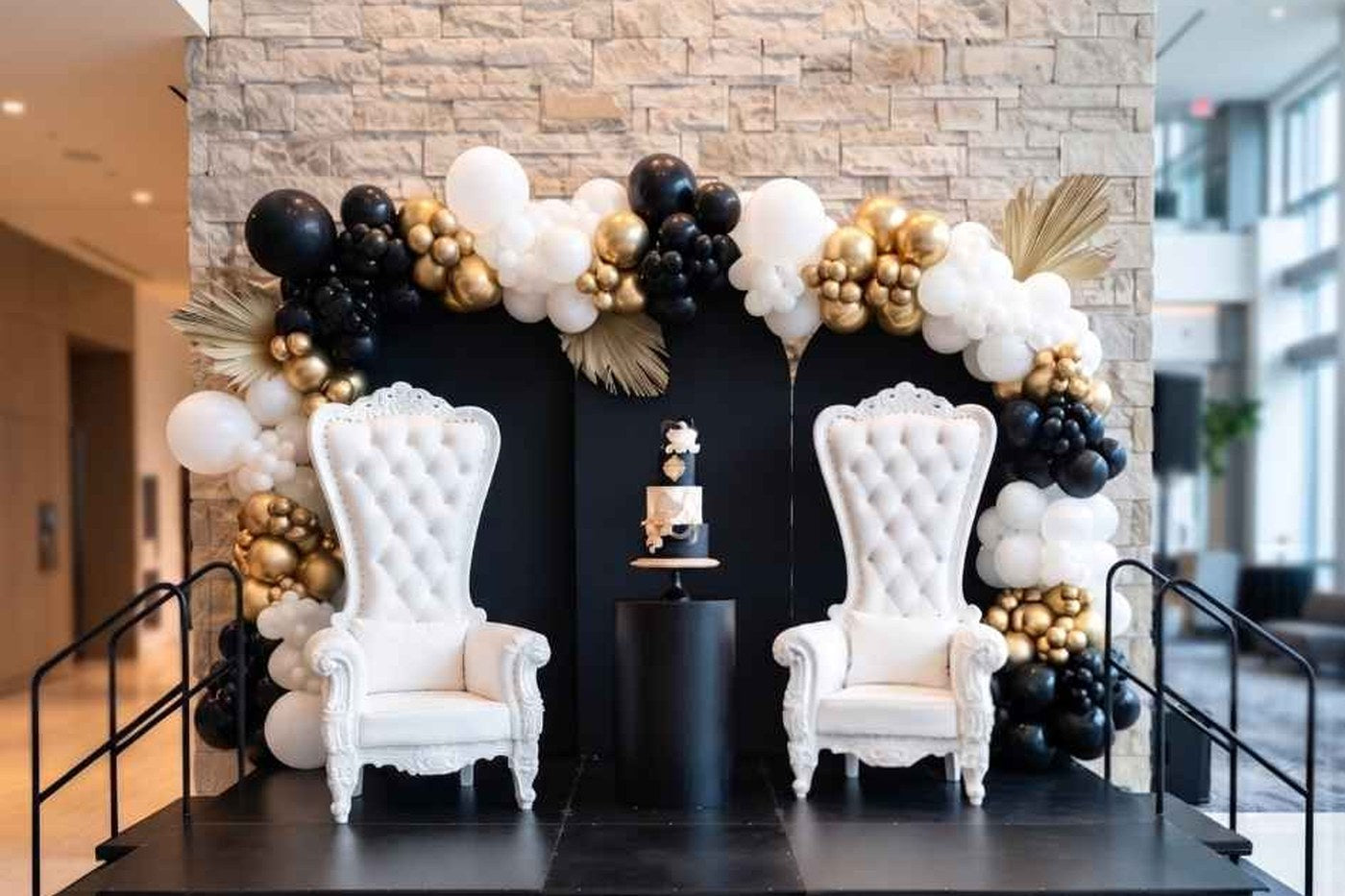 Two white throne chairs on a black-skirted portable stage under a black and gold balloon installation